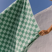 Green checkered beach towel being held against a blue sky and white wall.