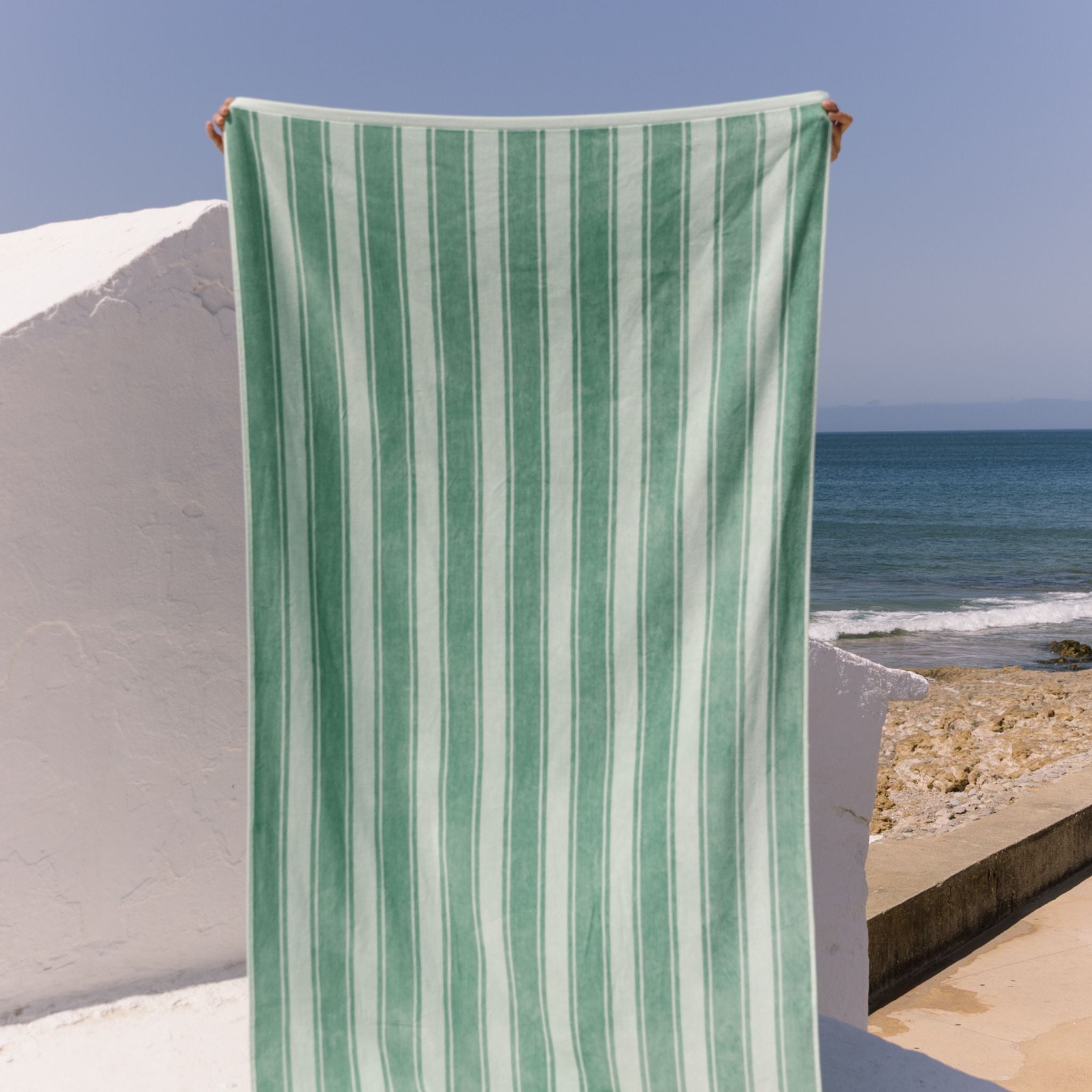 Green striped beach towel held up against a beach backdrop with ocean and sky.