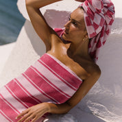Woman wrapped in a pink striped beach towel and with a checkered beach towel wrapped on her head, sitting on a white surface.
