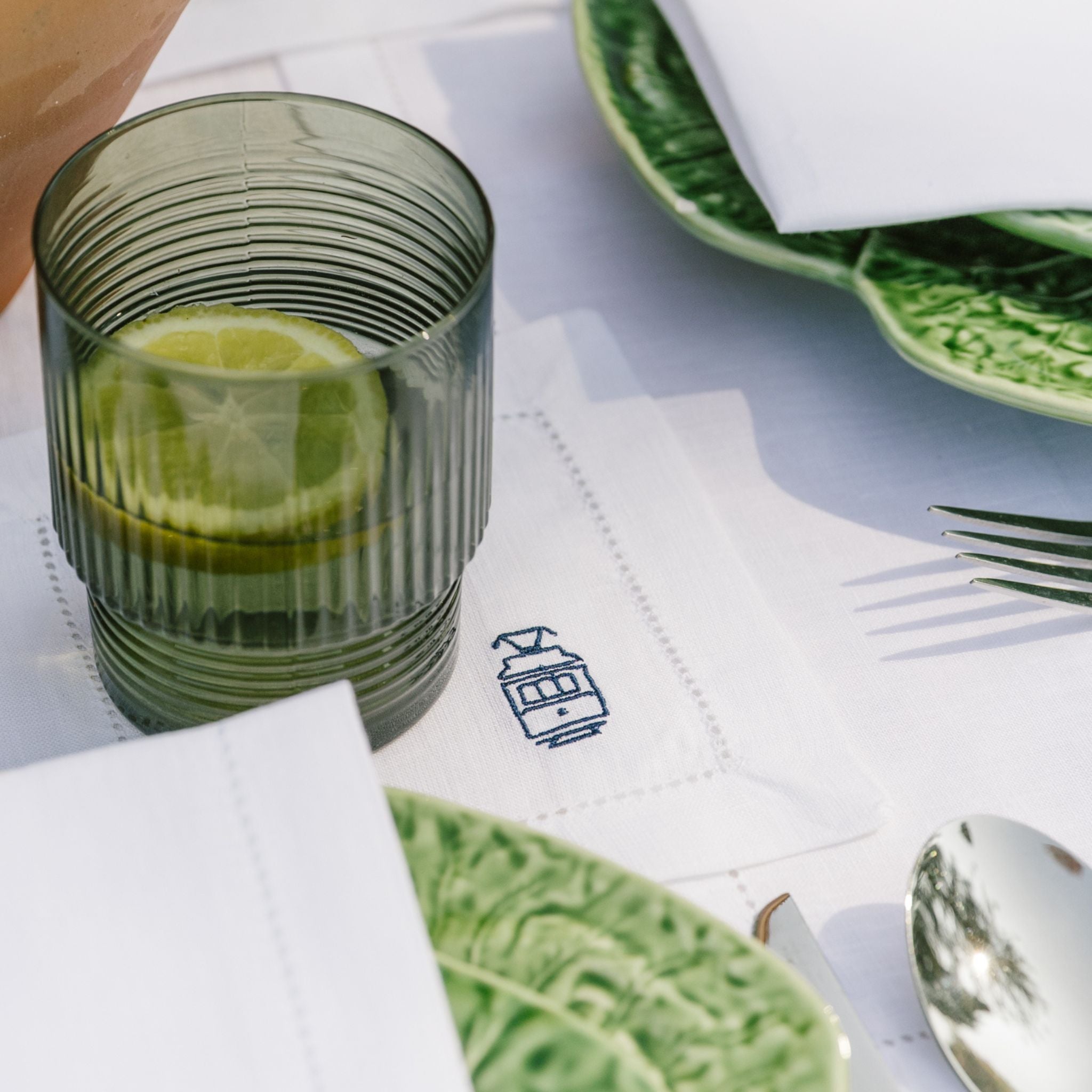 Green glass with lemon slices on a table setting with plates and cutlery.
