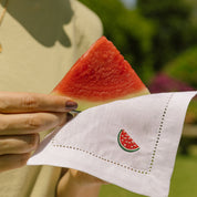 Person holding a slice of watermelon over a white napkin with an embroidered watermelon design.